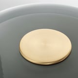 Un coperchio circolare color ottone in una ciotola arrotondata. Il coperchio è centrato e riflette la luce con una finitura liscia e lucida.