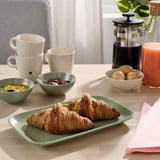 Un tavolo per la colazione con un piatto di croissant, ciotole di frutta e marmellata, tazze e una caffettiera, con le stoviglie FÄRGKLAR.