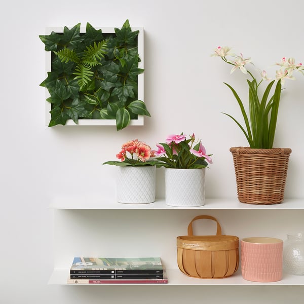 Mensole bianche con piante artificiali in vasi strutturati, un cesto di vimini con fiori e libri con un cesto di legno.