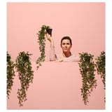 Una donna tiene una pianta finta rosa sopra la testa, circondata da piante artificiali appese dello stesso colore e design, contro un muro rosa.