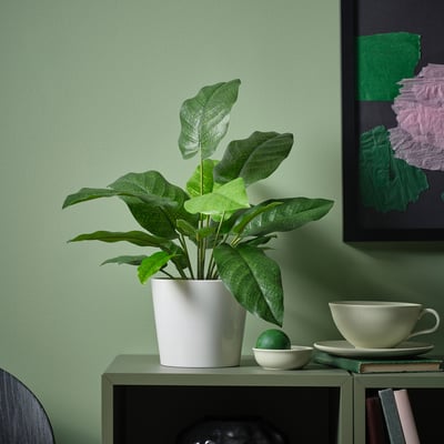 La piccola pianta verde FEJKA in vaso si trova su uno scaffale accanto a una tazza e a una ciotola, illuminando la stanza con foglie vivaci e realistiche.