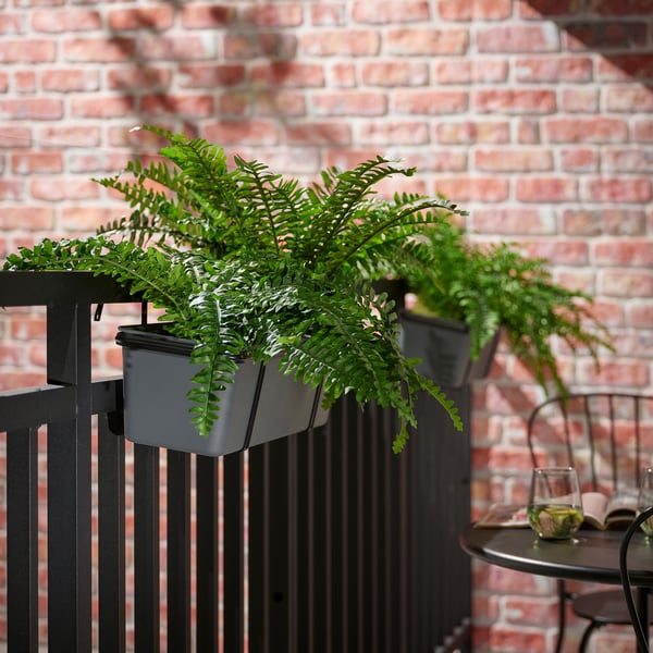 Ringhiera in metallo nero con piante artificiali FEJKA grigie, fogliame verde e un tavolino con vetro e libro.