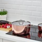 Una cucina moderna con pentole in acciaio inox e rame, pomodori e melanzane su un tagliere di legno accanto a un'erba bianca in vaso.