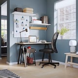 Home office con scrivania grigia, sedia nera, scaffali bianchi e laptop. Pareti, finestre e piante blu.