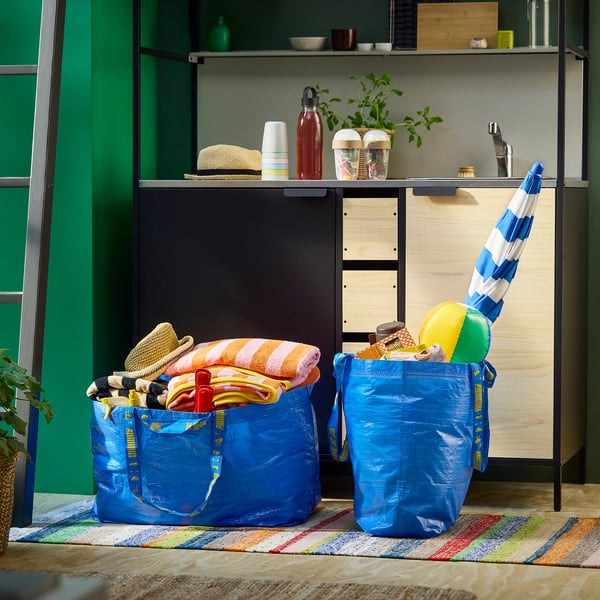 Due borse blu FRAKTA piene di asciugamani colorati e articoli da spiaggia sono posizionate su un tappeto a righe davanti a uno scaffale con oggetti da cucina.