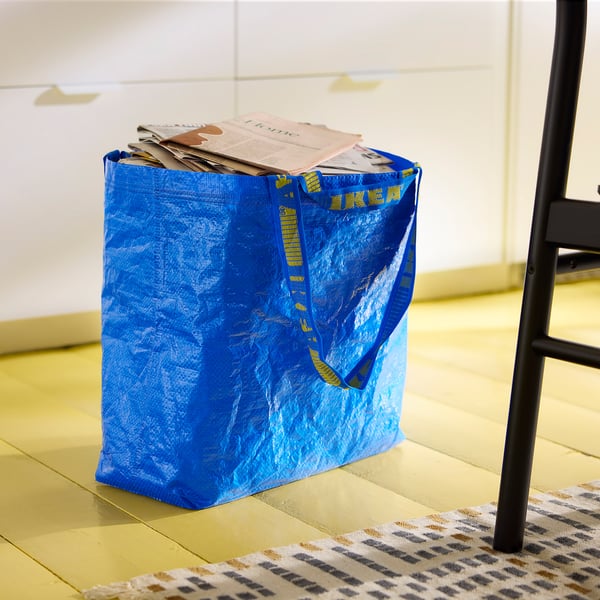 La borsa FRAKTA blu Ikea piena di carte si trova sul pavimento in legno. La borsa ha un testo giallo e due manici, uno lungo per il trasporto a spalla.