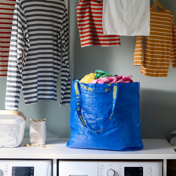 Borsa della spesa riutilizzabile blu, piena di vestiti colorati, seduta vicino a camicie a righe su grucce e due lavatrici.