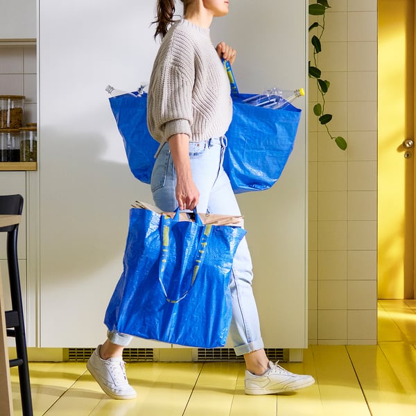 Persona che porta due grandi borse blu FRAKTA nel corridoio, resistenti e spaziose.