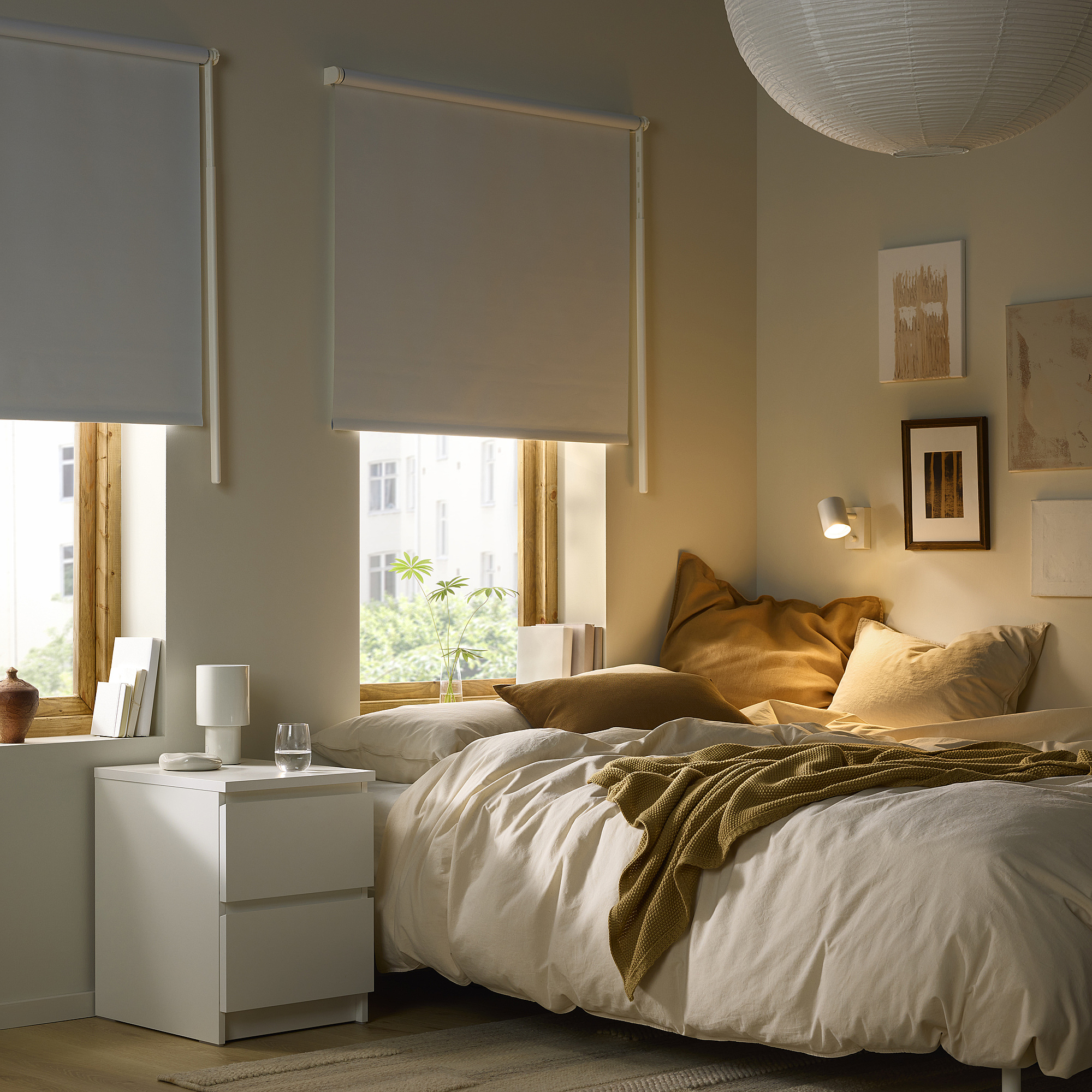 Accogliente camera da letto con pareti beige, persiane FRIDANS, letto pulito, comodino, lampada, libri e vetro. Luce naturale, lampada a sospensione.