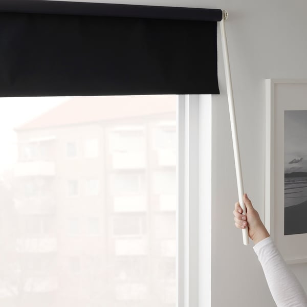 Persona che regola la tenda a rullo nera usando una bacchetta, bloccando la luce solare attraverso una finestra.