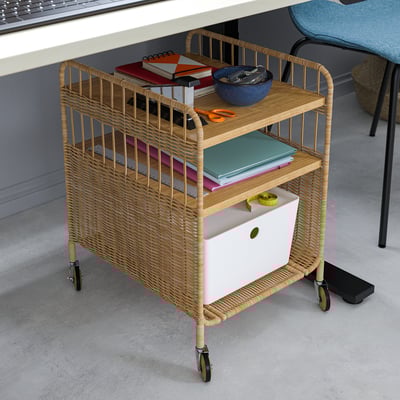 Carrello portaoggetti naturale con vimini intrecciato, ripiani in legno e ruote. Contiene libri, ciotole e forniture per ufficio.