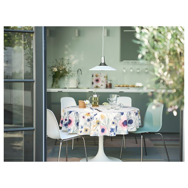 Scena da pranzo moderna con tavolo a tulipano bianco, tovaglia floreale colorata, sedie blu, lampada bianca sospesa, bicchieri e alzatina da cucina verde.