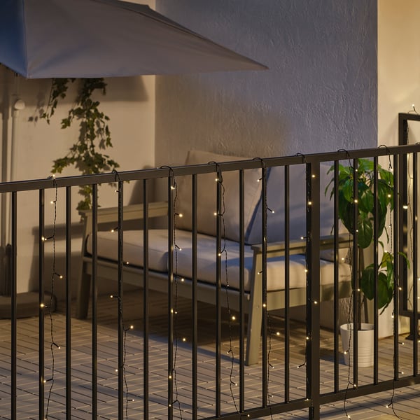 Balcone di notte con ringhiera infilata con lucine bianche calde. Sono visibili una panchina e piante in vaso.