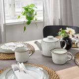 Set da tavola con set da tè bianco, comprensivo di teiera, tazze e brocca per crema. Tovagliette grigie e fiori in vaso nero. Luce naturale dalla finestra.