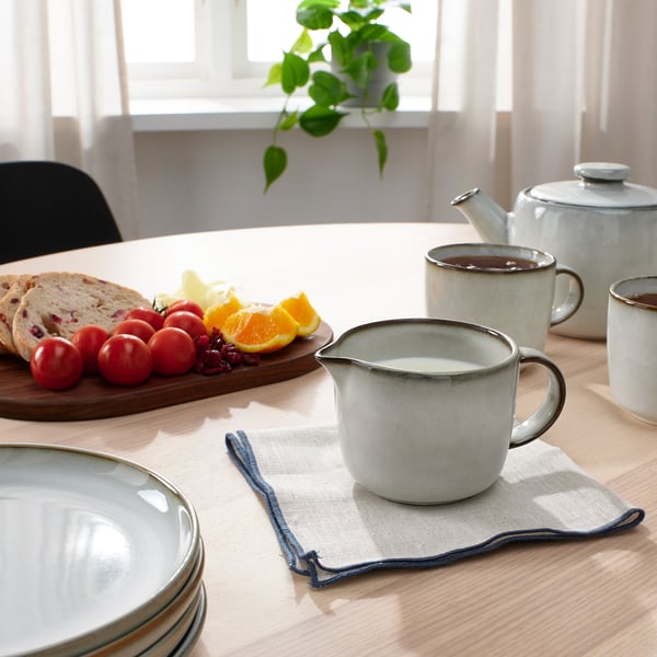 Tavolo da colazione con tazze GLADELIG, brocca per crema, frutta, pane, pianta.
