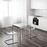 Cucina moderna con un piccolo tavolo con piano in vetro trasparente e due sedie trasparenti, vicino a armadi bianchi e un forno.