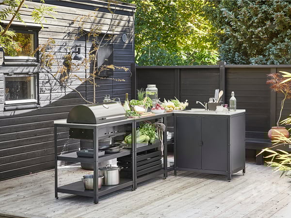 La cucina all'aperto ha una griglia, un lavello e ripiani in acciaio inossidabile. Recinzioni in legno scuro, verdure e utensili esposti.