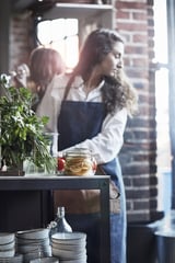 Donna in cucina, che indossa grembiule blu, che interagisce con il tavolo in acciaio inossidabile, tenendo il barattolo.