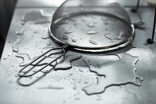 Utensili da cucina su superficie in acciaio inox bagnata. Il setaccio e le pinze sono visibili.