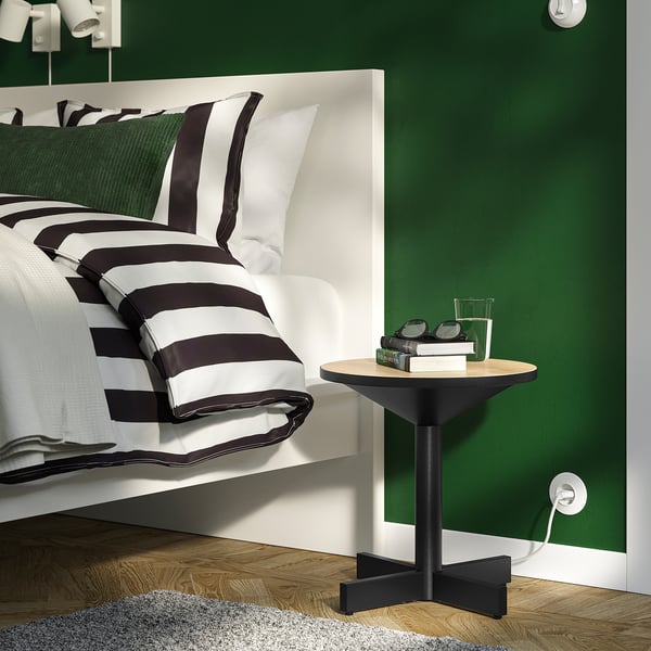 Piccolo e moderno comodino GRIMSARBO contro la parete verde accanto al letto bianco con coperta a righe bianche e nere.