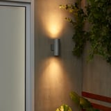 Lampada da parete moderna per esterni grigia GRÖNSPRÖT su parete beige vicino alla porta bianca, con luce calda e contrasto di verde.