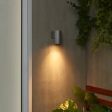 La piccola lampada da parete nera GRÖNSPRÖT emette una luce calda accanto alla porta bianca, circondata da piante verdi, evidenziando le sue dimensioni compatte e il design elegante.