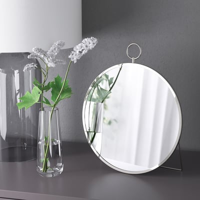 Uno specchio rotondo su un supporto accanto a un vaso trasparente con fiori bianchi. Lo specchio può essere appeso o in piedi.