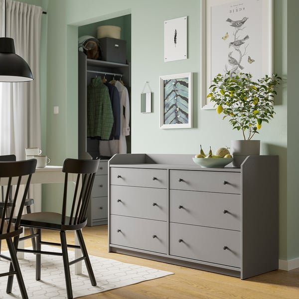 Interni della casa con comò grigio HAUGA, arte, piante, sedie nere, tazze bianche, armadio con vestiti, tende.
