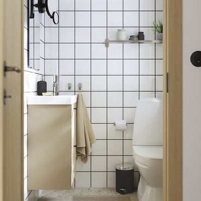 Un bagno minimalista con piastrelle bianche a griglia, un mobile beige opaco, un lavandino e servizi igienici bianchi e una porta in vetro.