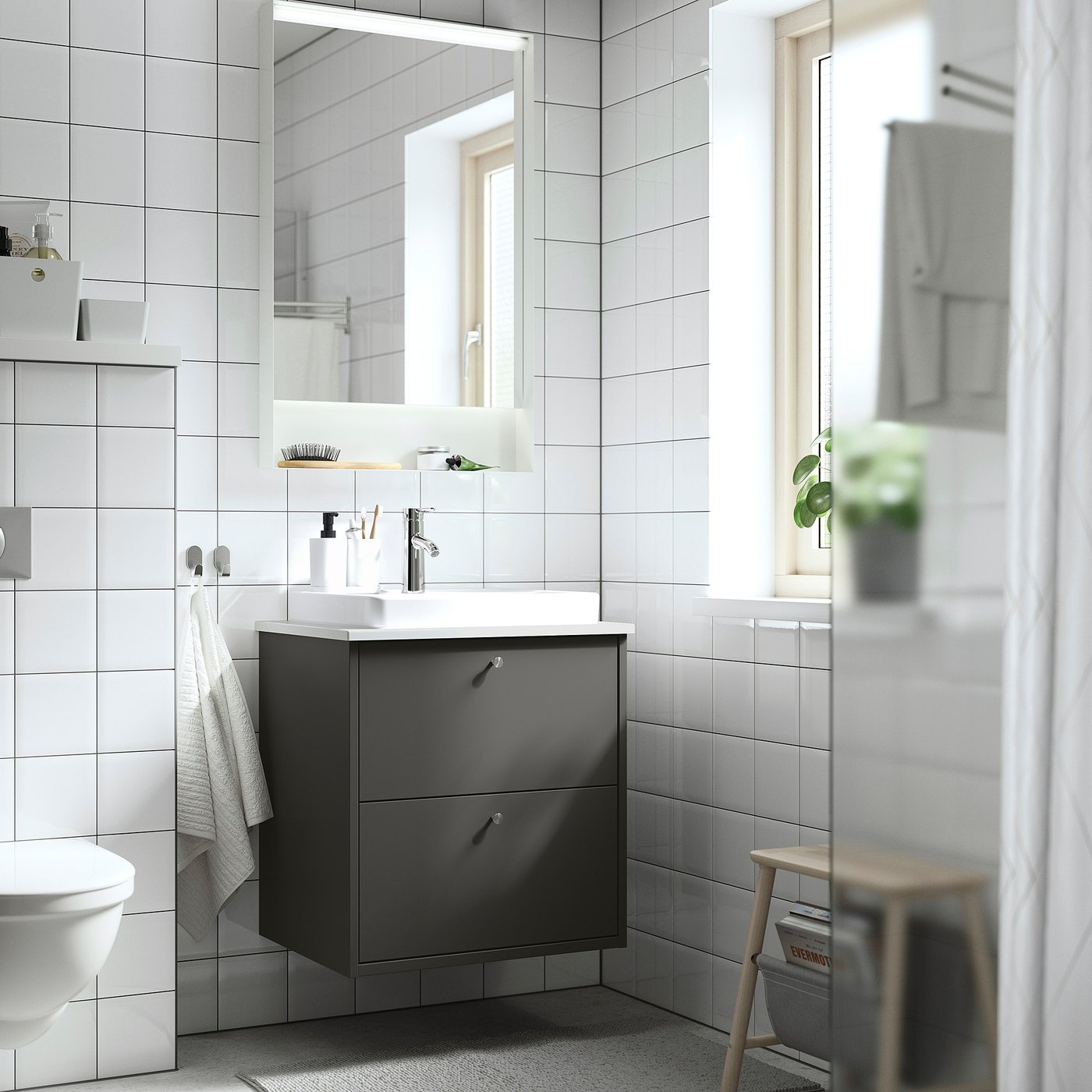 Bagno moderno con lavabo HAVBÄCK grigio scuro, lavandino bianco, grande specchio e cassetti portaoggetti. Le pareti piastrellate bianche e lo sgabello in legno aggiungono calore.