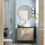 Bagno moderno con lavabo HAVBÄCK beige, maniglie nere, lavandino bianco, doccia in vetro, pareti piastrellate blu, specchio rotondo e pianta verde.