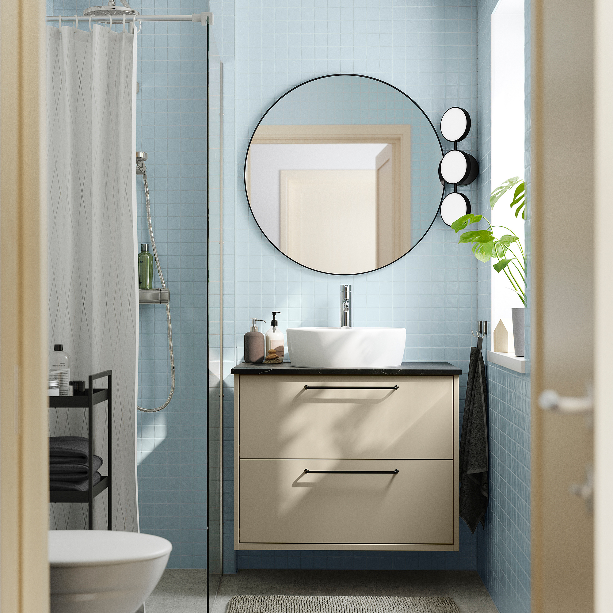 Bagno moderno con lavabo HAVBÄCK beige, maniglie nere, lavandino bianco, doccia in vetro, pareti piastrellate blu, specchio rotondo e pianta verde.
