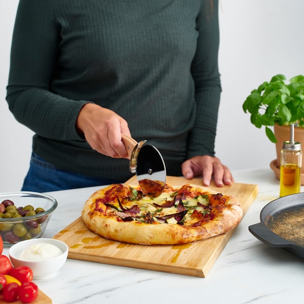 Persona che affetta la pizza fatta in casa sul tagliere con tagliapizza; circondato da condimenti e salsa.