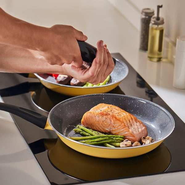 Persona che condisce il salmone in una padella antiaderente gialla con asparagi verdi. Due pentole che cuociono su un fornello.