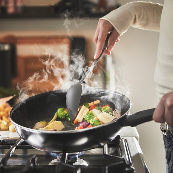 Persona che cucina le verdure in una padella antiaderente su un fornello, vapore in aumento.