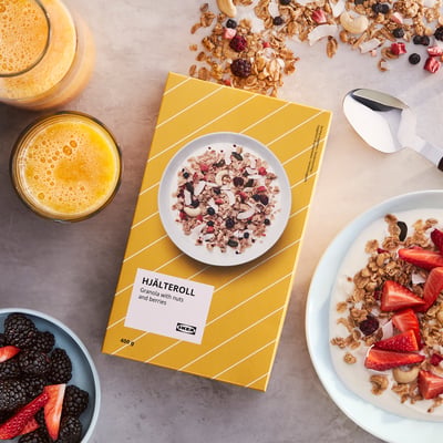 Una ciotola di muesli HJÄLTEROLL con noci e frutti di bosco su un tavolo con succo d'arancia e yogurt. La confezione è aperta, mostrando la granola all'interno.
