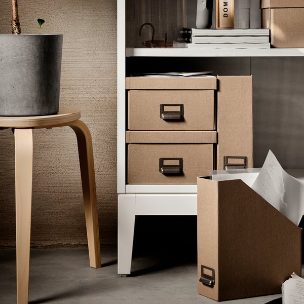 Presenta un moderno mobile contenitore IDANÄS bianco con cassetti in texture naturale e una fioriera in cemento su uno sgabello in legno.