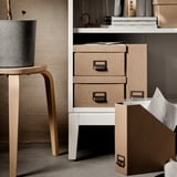 Presenta un moderno mobile contenitore IDANÄS bianco con cassetti in texture naturale e una fioriera in cemento su uno sgabello in legno.