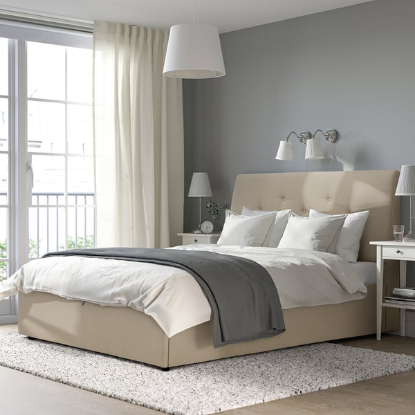 Pouf letto IDANÄS beige sotto una grande finestra, vestito di lenzuola bianche, accanto a un comodino con lampade e libri. Pareti grigie, tappeto accogliente.