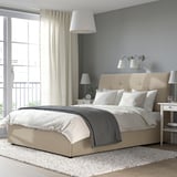 Pouf letto IDANÄS beige sotto una grande finestra, vestito di lenzuola bianche, accanto a un comodino con lampade e libri. Pareti grigie, tappeto accogliente.