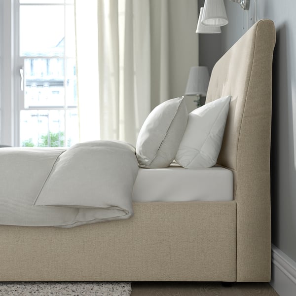 Un letto imbottito beige con testiera inclinata, dotato di morbidi cuscini e una coperta piegata, che offre un comodo supporto e spazio di archiviazione.