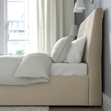 Un letto imbottito beige con testiera inclinata, dotato di morbidi cuscini e una coperta piegata, che offre un comodo supporto e spazio di archiviazione.