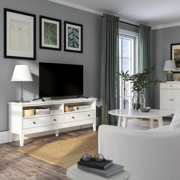 Soggiorno moderno con porta tv IDANÄS bianco, tv a schermo piatto, lampada, tavolo rotondo, sedia, tappeto grigio e tende verdi. Opera d'arte su pareti grigio chiaro.