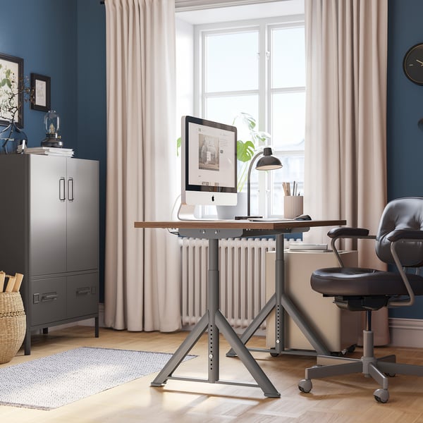 Configurazione dell'home office con scrivania IDÅSEN, parete blu, gestione dei cavi, sedia grigia, armadio, cestino, quadri e pianta.