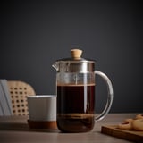 La french press in vetro trasparente con coperchio e manico in legno si trova su un tavolo accanto a una tazza bianca e un piatto di biscotti.