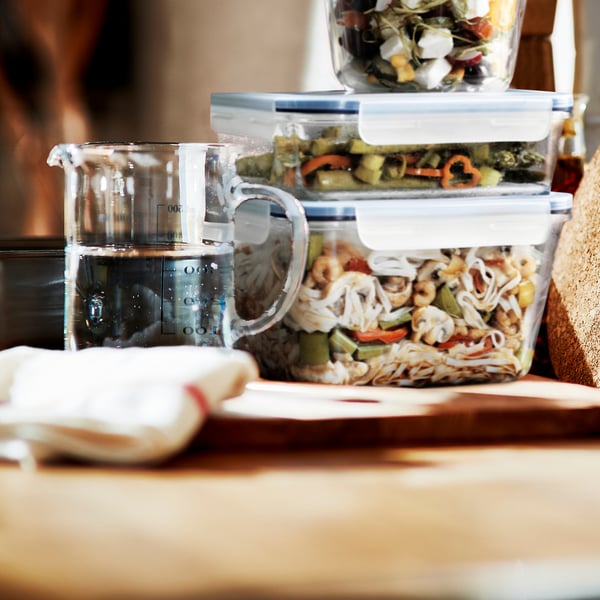 Contenitori di vetro impilati con coperchi a serratura, uno contenente verdure e noodles, posizionati su un tavolo di legno accanto a una brocca dosatrice d'acqua.