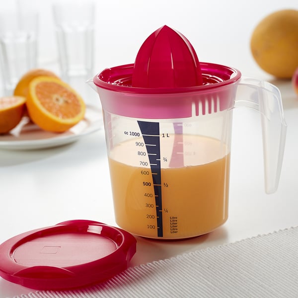 Spremiagrumi rosa con brocca trasparente piena di succo d'arancia, che mostra segni di misura fino a 1 litro. Il coperchio si trova accanto ad esso.