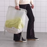 Persona che tiene un sacco portabiancheria leggero e pieghevole con vestiti gialli, contro il muro piastrellato bianco.