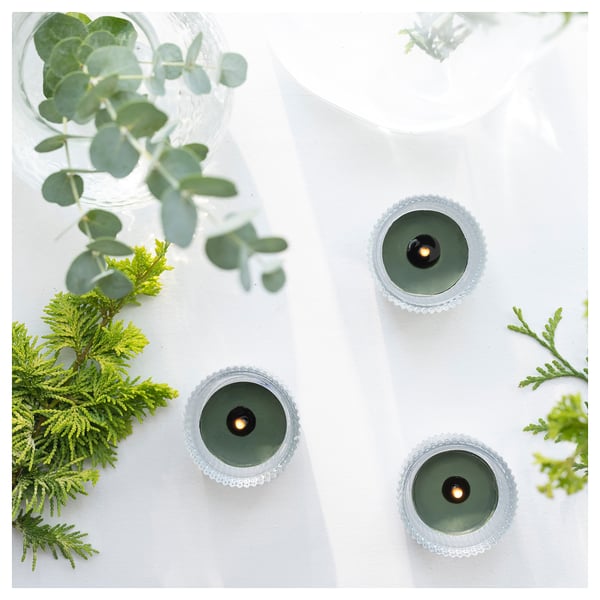 Tre candele verdi con portacandele intrecciate, circondate dal verde e dalle foglie di eucalipto su sfondo bianco, che emettono un bagliore caldo e invitante.