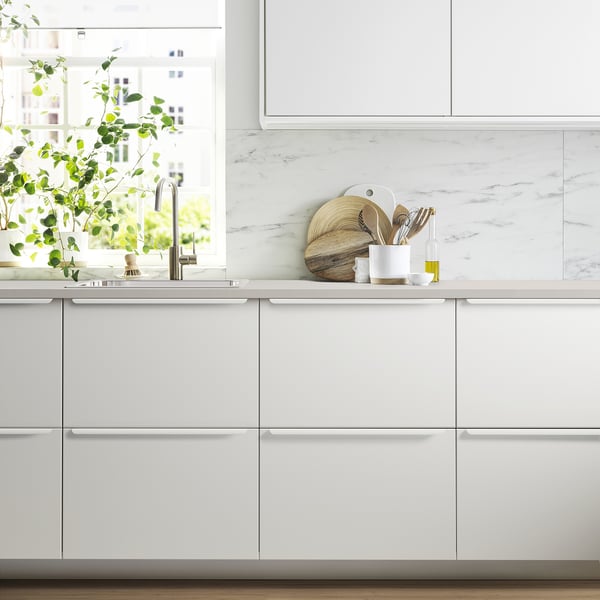 Cucina moderna bianca: piano di lavoro JÄRSTORP, alzatina in marmo, utensili in legno, pianta in vaso. Lavello e rubinetto visibili.